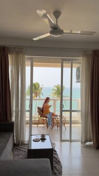Interior living room writer caucasian woman sitting at balcony with notebook, warm light filtering through curtains, palm trees