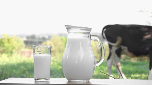 A glass and jug filled with fresh milk are placed on a wooden surface, set against a rural landscape with a cow in the background. The scene highlights organic farming and wholesome dairy products