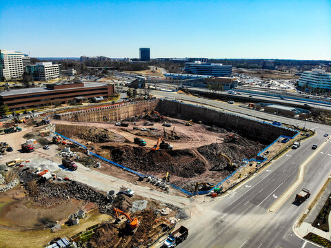 Reston, Virginia, United States - 12 March 2019: Aerial view of a construction site with excavators and dump trucks in a deep pit, surrounded by office buildings and a highway.