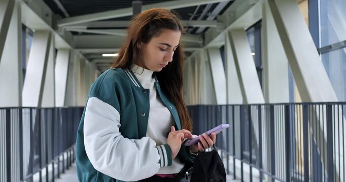 Young woman typing message closely in glass corridor, closeup hands and phone, serious concentration for caption editing, modern beams and railings provide clean background for social content creation