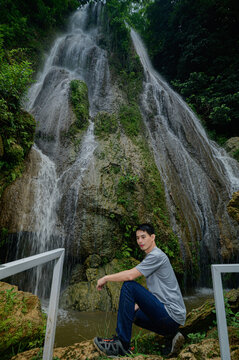 Young man sitting near large cascading waterfall in lush green forest landscape