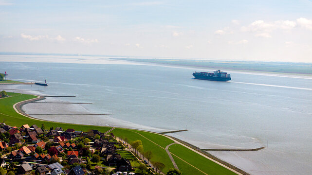 Brunsbuttel, Schleswig-Holstein, Germany - 30 May 2025: Aerial view of a large container ship navigating the Elbe river near the green dikes and residential houses of Brunsbuttel.