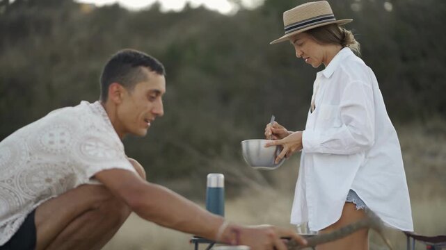 A man crouches by a campfire, tending to a pot, as a woman mixes ingredients in the background on a sandy beach. The scene highlights outdoor cooking, travel, and summer relaxation