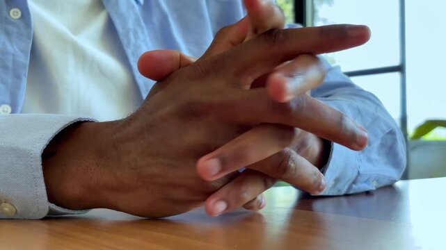 A close-up shot captures an African American man's hands fidgeting nervously on a wooden table. His fingers interlace and rub, conveying a sense of anxiety, contemplation, or quiet waiting.