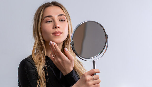 Woman focused on skincare, anti-ageing skin care. Young woman face examining skin with mirror concentrated closeup. Dermatology. Beautiful woman looking in the mirror, touching her face with her hand