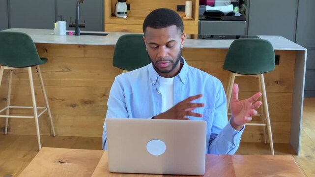 A young Black man uses his laptop for a video call or remote work from his modern home kitchen. He gestures actively, conveying communication and engagement in a contemporary setting.