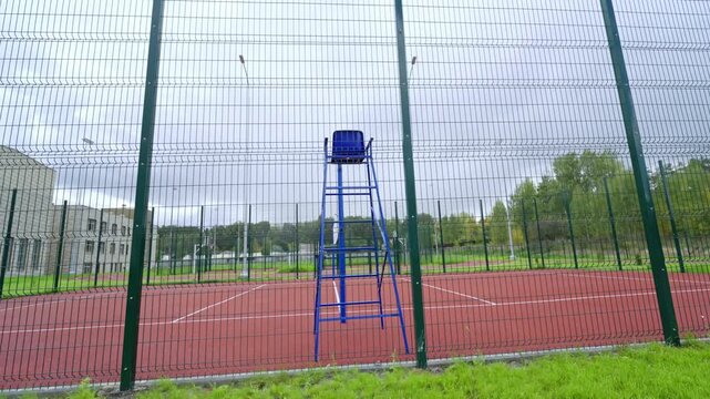 Referee chair stands beside volleyball net on red court. High metal fence surrounds professional sports area under cloudy autumn sky