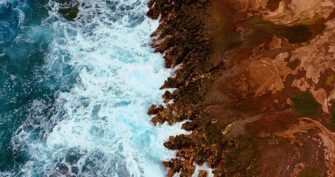 Turquoise ocean wave crashing on lava rock shoreline, overhead view, Hawaii. White foam surf meeting jagged dark volcanic rock, rust and brown coastal surface with patches of algae.