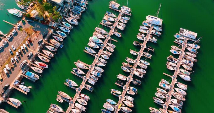 Sailboat and yacht marina docks. Overhead drone perspective showing parallel finger piers over bright green water, rows of white motorboat and sailboat, parking lot and palm tree.