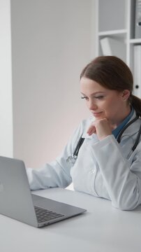 Confident female physician using laptop for telemedicine consultation or electronic health records management in bright clinic. Modern healthcare technology and digital workflow concept