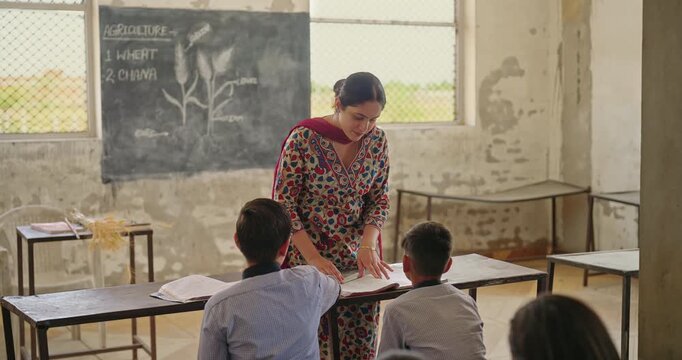 Indian young adult lady female teach lesson do study work indoor class room rural area place. Teen boy child sitting on bench wear uniform look tutor talk speak share story learn enjoy day desi life