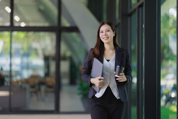 Fototapeta premium Young Asian businesswoman walking through a bright office hallway, carrying a laptop and coffee, symbolizing a dynamic professional lifestyle and entrepreneurial spirit