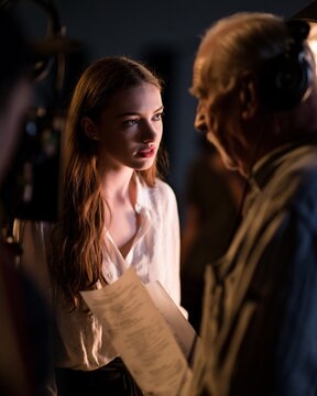 A young actress and an older actor exchange a tense look on a dimly lit film set, with soft key light, shallow depth of field, script pages, and a camera rig creating a dramatic behind-the-scenes