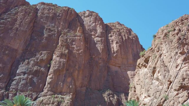 In Morocco's Todra Gorge, towering cliffs and rugged rock formations rise above an arid desert landscape accented by scattered palm trees and vegetation.