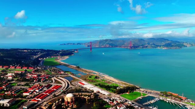 Aerial view of Golden Gate Bridge and Presidio shoreline, San Francisco, California. Drone perspective over red roofed fort buildings, sandy cove, turquoise bay and Marin hills under blue sky.
