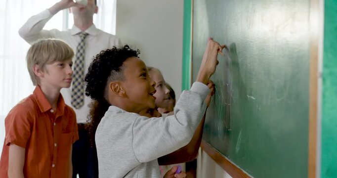Multicultural elementary pupil group writes math answer on green chalkboard while adult male teacher guides, building back to school participation, teamwork, confidence, positive classroom routine.