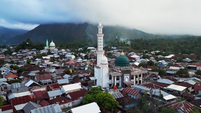 Mosques rising above a mountain village surrounded by rural homes and cloud covered peaks in Sembalun Lombok, aerial drone footage.