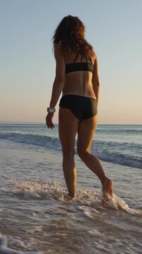 Rear view of a young woman strolling on the shoreline, her feet touching the gentle surf. Serene beach scene captures relaxation, freedom, and the beauty of a summer sunset by the water