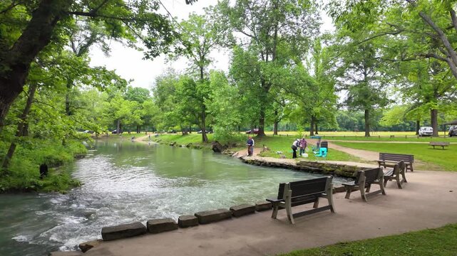 View of Maramec Spring Park with people fishing along the riverbank, wooden benches, and lush green trees in Saint James, Missouri, United States.