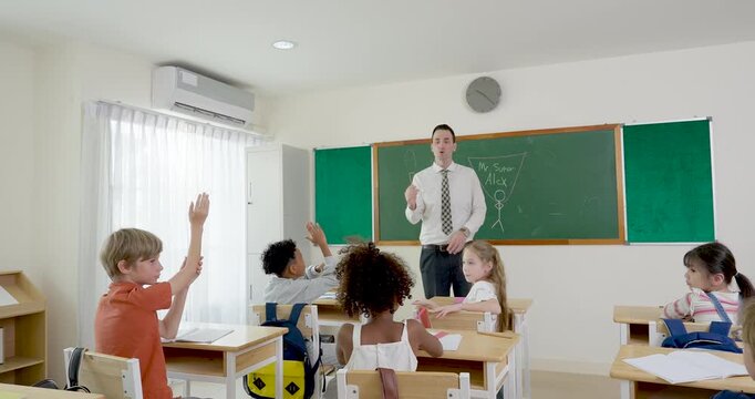 Multicultural elementary pupil group raises arms during math question time as male teacher guides lesson at chalkboard, interactive classroom routine builds confidence, attention, motivation