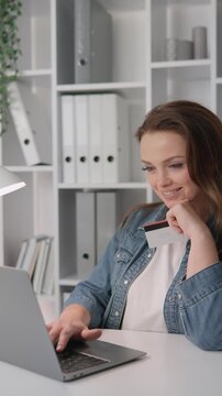 Confident young woman makes an online purchase using her credit card and laptop in a bright office setting. E commerce, remote work, and digital finance trends are highlighted in this scene