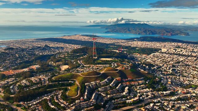 Sutro Tower above San Francisco western neighborhoods toward Golden Gate. Drone shot of red lattice antenna tower on forested hill, bay and Marin County mountains visible at horizon under blue sky.