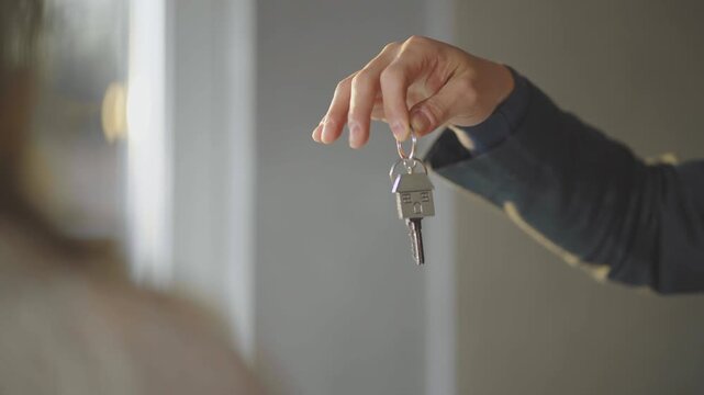 A man's hand holds up a house key with a metal house-shaped keychain and passes it to an unidentifiable woman.