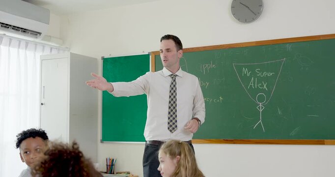 Male adult teacher points across elementary classroom while giving instruction near chalkboard, guiding diverse student group through lesson notes, question time, classroom rule and learning routine