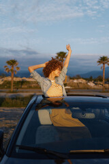 Fototapeta premium Young woman with curly hair leans out of a car window, arms raised in a carefree moment under a bright blue sky on a scenic roadside drive with warm sunlight and distant hills.