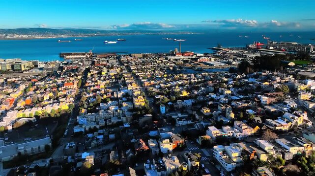 Colorful Dogpatch and Potrero Hill rooftops toward Oakland across bay. Wide aerial view of multicolored house grid, industrial waterfront, cargo ship on deep blue water and East Bay hills.