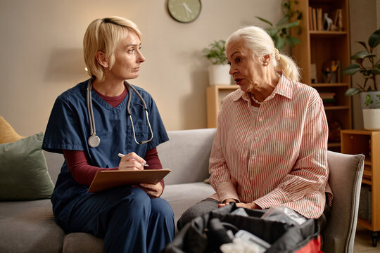 Medium shot of Caucasian nurse listening and taking notes while senior woman speaking during home care visit, discussing health concerns. Useful for eldercare, patient support, caregiving marketing
