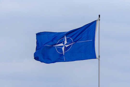 Die blaue NATO-Flagge mit dem charakteristischen wei&szlig;en Kompassstern weht im Wind an einem Fahnenmast vor bedecktem Himmel.