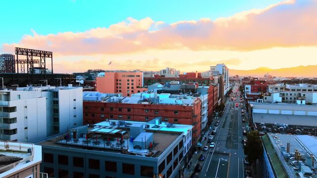 Mission Bay neighborhood street view at dusk, San Francisco. Baseball stadium truss at left, brick warehouse row, straight boulevard receding toward mountain horizon under peach and teal sky.