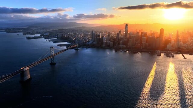 Sunset over downtown skyline and bay bridge, San Francisco. Golden sunburst above financial towers, Bay Bridge suspension span crossing dark water and mountain silhouette on horizon.