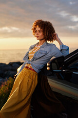 Fototapeta premium Young woman leans against a car at sunset by the coast, wearing a striped top, denim shirt and flowing mustard skirt, feeling contemplative and relaxed on a warm evening by the sea.