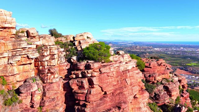 Rocky cliff edge overlooking coastal plain and Mediterranean sea, Spain. Side view of red sandstone ledge with green tree, patchwork farmland, coastal city and blue ocean horizon under clear sky.