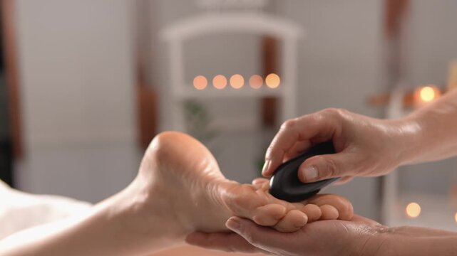 Close up of hands performing a hot stone massage on a person's foot in a cozy spa setting with blurred candles in the background. The scene evokes calmness, self care, and rejuvenation