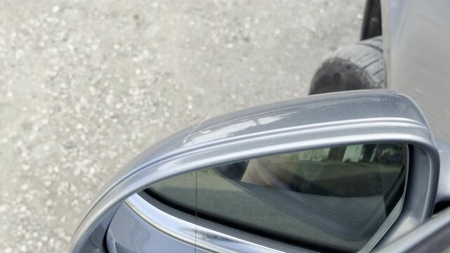 An automatic car mirror folds tightly against the vehicle on a gravel path. The motorized mechanism produces a low whirring and clicking sound.