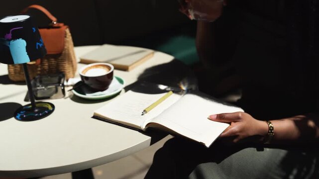 A black woman writes in a notebook paper diary closeup in a cafe on a sunny day outdoors