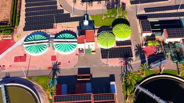 Covered digester and solar panel array at treatment plant, Spain. Top-down perspective of teal and green scalloped tank cover, palm tree shadow, photovoltaic roof and orange paved yard.