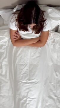 A young woman lies on a bed, experiencing abdominal discomfort, possibly period pain or cramps. Her arms are crossed over her stomach, conveying distress and unease in a neutral setting.