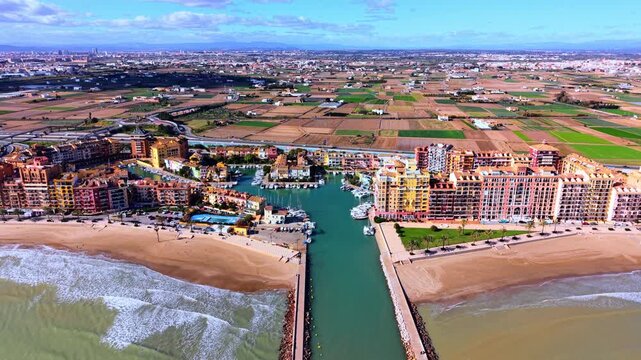 Marina and colorful waterfront apartments, Valencia region, Spain. Inland over teal canal, moored sailboat and motorboat, sandy beach and patchwork agricultural plain stretching to city skyline.