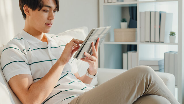 Young Asia male with casual cloth sit on sofa finger scrolling digital tablet favorite online movie in living room at cozy house. E-book reading, shopping online, Lifestyle leisure at home concept.