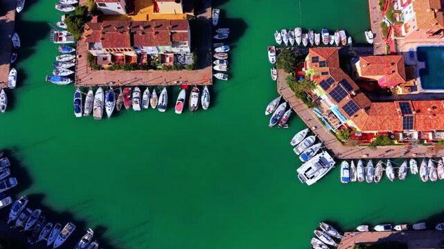 Overhead view of marina boats moored in green canal water, Valencia, Spain. Rows of sailboat and motorboat at dock, terracotta rooftop, solar panel, and vivid emerald water channel.