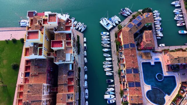 Marina canal flanked by terracotta rooftops and moored boats, Valencia, Spain. Teal waterway, yellow and orange residential building with pool, and dense vessel row along quayside.