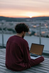 Obraz premium Man on rooftop working on laptop at sunset with city and hills in the distance for background and banner use