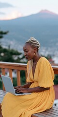 Obraz premium Young woman in yellow dress working on a laptop on a wooden balcony at sunset with mountain and city view for remote work background and lifestyle design