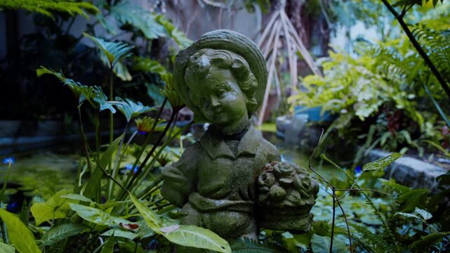 植物園の池のほとりに佇む苔むした子供の石像と緑豊かな庭園の風景 4Kシネマティック映像素材