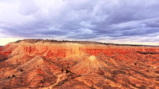 Red sandstone mesa and badland landscape under stormy sky. Wide drone perspective of flat topped cliff face with orange and ochre rock layer, eroded gully, sparse conifer, dirt track.