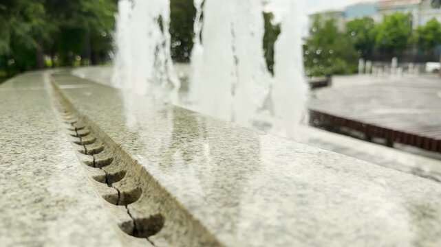 A polished granite fountain ledge reflects the light. Water jets spray upward in the background. The fountain is located in an open city park.